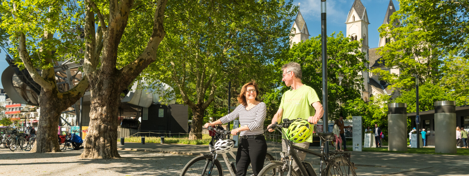 Pärchen mit E-Bikes lächeln. Seilbahn-Talstation und Basilika St. Kastor sind im Hintergrund zu sehen. ©Koblenz-Touristik GmbH, Dominik Ketz