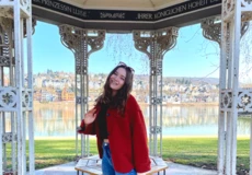Frau mit rotem Mantel und Sonnenbrille steht im Augusta-Pavillon in den Rheinanlagen, im Hintergrund der Rhein und der Stadtteil Pfaffendorf.