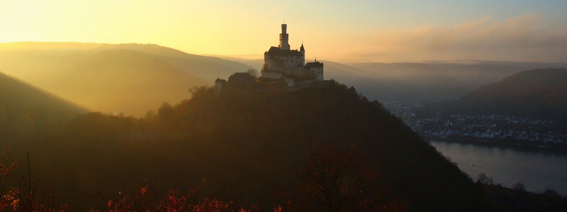 Die Marksburg auf einer Höhe mit Rhein und benebelten Bergen beim Sonnenuntergang ©