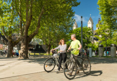 Pärchen mit E-Bikes lächeln. Seilbahn-Talstation und Basilika St. Kastor sind im Hintergrund zu sehen. ©Koblenz-Touristik GmbH, Dominik Ketz