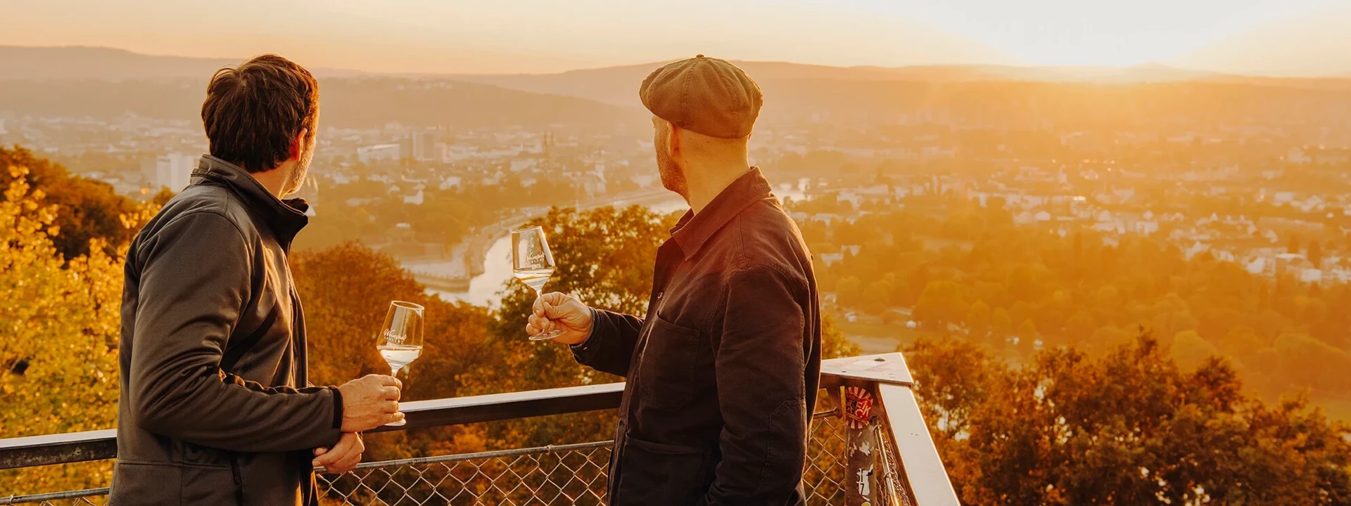 Zwei Männer von der Seite halten Weingläser und blicken auf die Stadt Koblenz beim Sonnenuntergang ©Koblenz-Touristik GmbH, Philip Bruederle