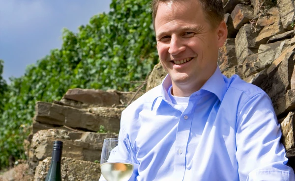 Betreiber des Weingut Lunnebach lächelt mit einem Glas Weißwein in der Hand und seinem Weinberg im Hintergrund