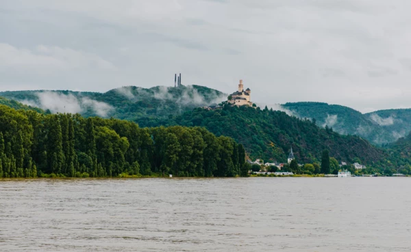 Blick auf die Marksburg vom Rheinufer umgeben von Bergen und Wald mit dem Rhein im Vordergrund