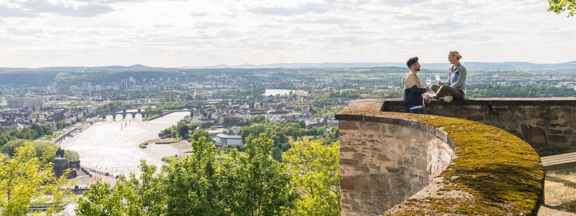 Pärchen sitzt auf der Mauer der Festung Ehrenbreitstein und hält Weingläser mit Stadt Koblenz im Hintergrund ©Koblenz-Touristik GmbH, Dominik Ketz