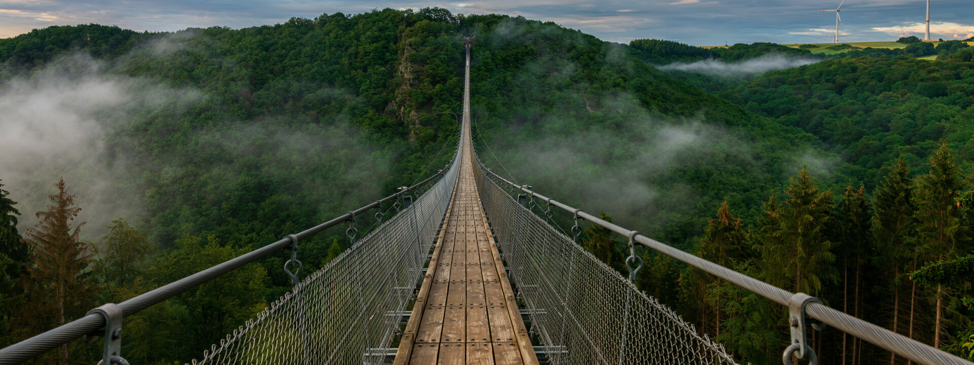 Blick auf eine lange Hängeseilbrücke, die sich über ein tiefes bewaldetes Tal erhebt, im Hintergrund bewölkter Himmel und Nebelschwaden im Tal  ©Bernhard - stock.adobe.com