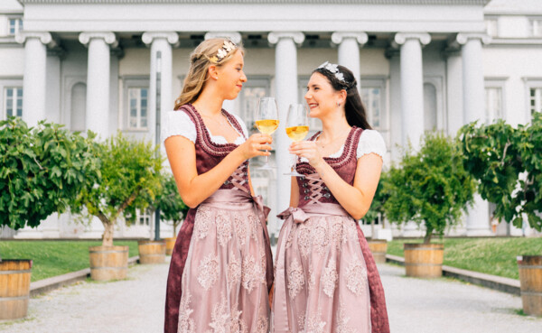 Zwei junge Frauen in festlichen Kleidern, mit Diadem und Weingläsern in der Hand, im Hintergrund das kurfürstliche Schloss in Koblenz ©Koblenz-Touristik GmbH, Tibor Resch