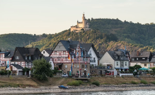 Blick auf die Marksburg, umgeben von Wald, im Vordergrund der Rhein und Fachwerkhäuser