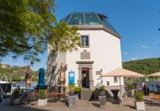 Das Pegelhaus in Koblenz mit Restauranttischen im Vordergrund und dem Rhein und der Festung Ehrenbreitstein im Hintergrund