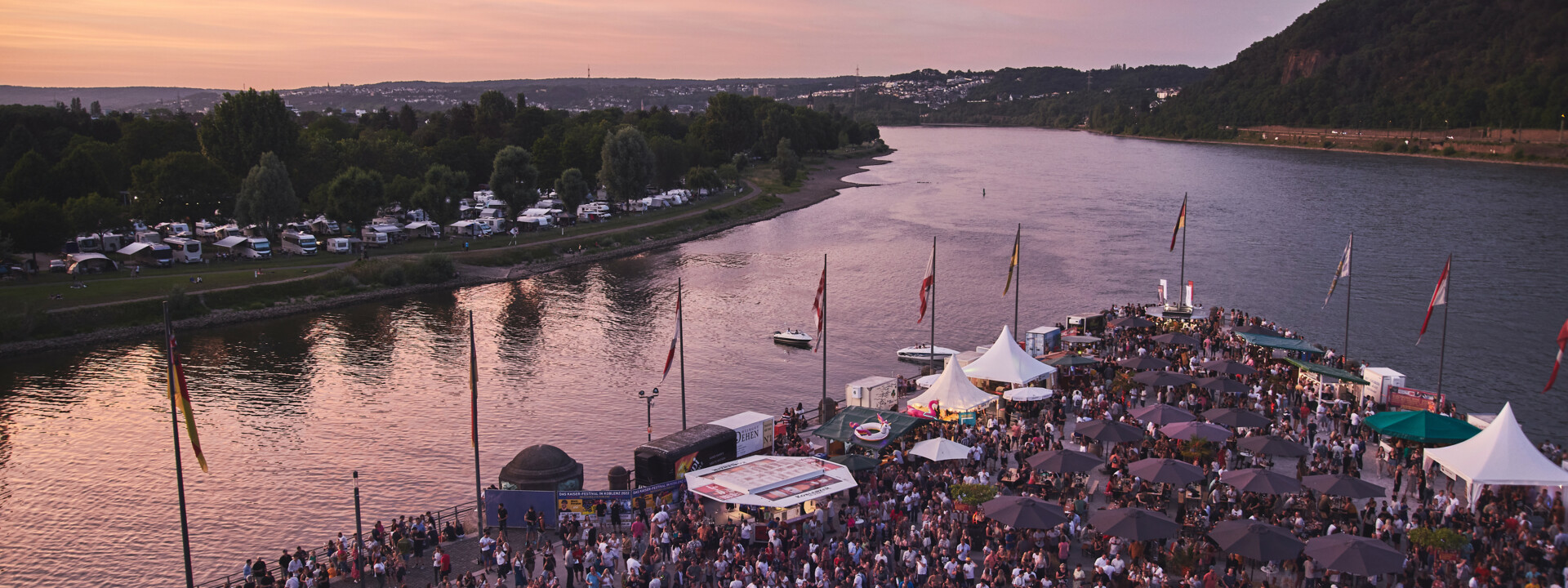 Blick von oben auf das Deutsche Eck bei Abenddämmerung während einer Veranstaltung, Menschengruppen stehen vor einer Bühne und an Ständen verteilt auf dem Gelände ©Picture Colada