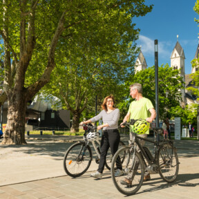 Pärchen mit E-Bikes lächeln. Seilbahn-Talstation und Basilika St. Kastor sind im Hintergrund zu sehen. © Koblenz-Touristik GmbH, Dominik Ketz
