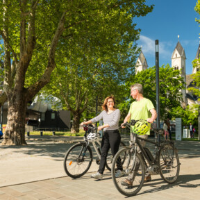 Pärchen mit E-Bikes lächeln. Seilbahn-Talstation und Basilika St. Kastor sind im Hintergrund zu sehen. ©Koblenz-Touristik GmbH, Dominik Ketz