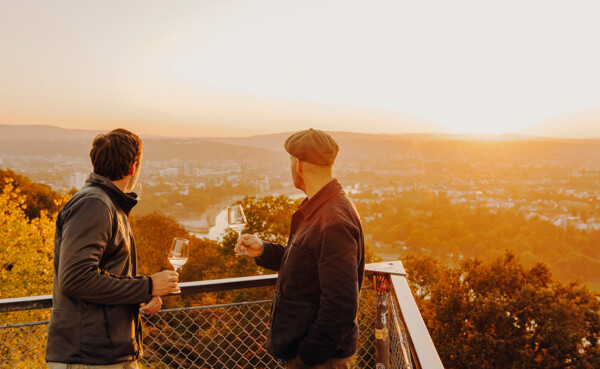 Zwei Männer im Abendlicht mit Weingläsern auf einer Aussichtsplattform mit Blick auf den Zusammenfluss von Rhein und Mosel ©Koblenz-Touristik GmbH, Philipp Bruederle