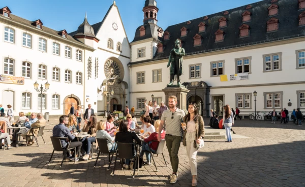 Pärchen von Vorne geht und Menschengruppen sitzen an Cafétischen auf dem Jesuitenplatz in der Koblenzer Altstadt