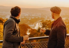 Zwei M&auml;nner von der Seite halten Weingl&auml;ser und blicken auf die Stadt Koblenz beim Sonnenuntergang &copy; Koblenz-Touristik GmbH, Philip Bruederle