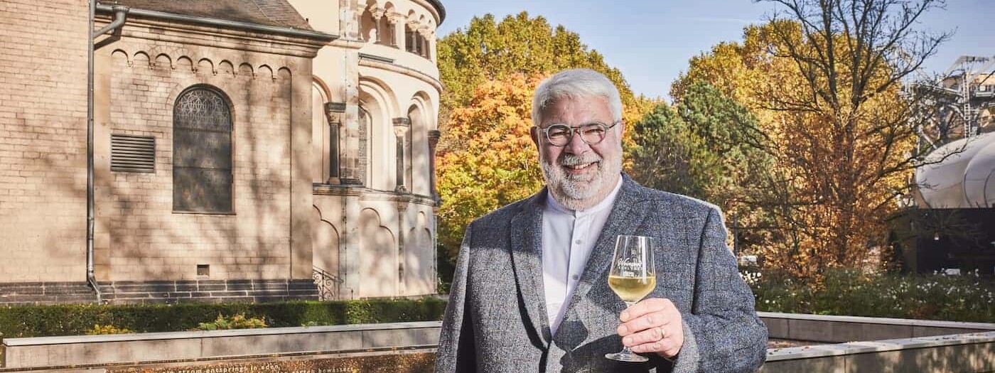 Lächelnder Stadtführer Michael Hörter mit einem Glas Wein in der Hand steht vor dem Basilika St. Kastor im Herbst ©Koblenz-Touristik GmbH, Picture Colada