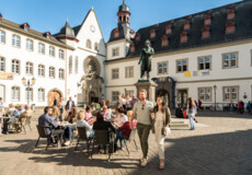 Pärchen im Vordergrund spaziert und Menschengruppen im Hintergrund sitzen an Cafétischen auf dem Jesuitenplatz in der Koblenzer Altstadt ©Koblenz-Touristik GmbH, Dominik Ketz