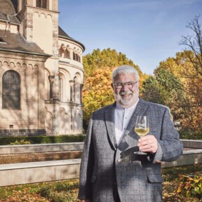 Lächelnder Stadtführer Michael Hörter mit einem Glas Wein in der Hand steht vor dem Basilika St. Kastor im Herbst © Koblenz-Touristik GmbH, Picture Colada
