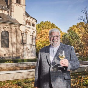 Lächelnder Stadtführer Michael Hörter mit einem Glas Wein in der Hand steht vor dem Basilika St. Kastor im Herbst ©Koblenz-Touristik GmbH, Picture Colada