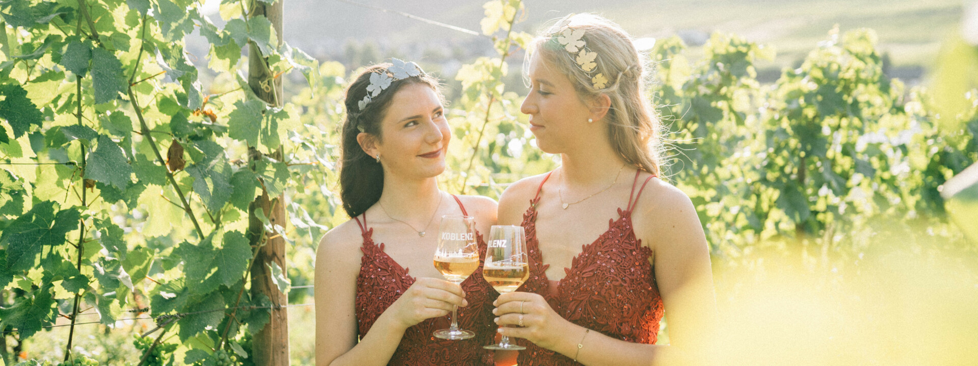 Zwei junge Frauen mit roten Festkleidern, Diadem und Weingläsern, im Hintergrund Weinberge ©Koblenz-Touristik GmbH, Tibor Resch