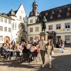 Pärchen im Vordergrund spaziert und Menschengruppen im Hintergrund sitzen an Cafétischen auf dem Jesuitenplatz in der Koblenzer Altstadt © Koblenz-Touristik GmbH, Dominik Ketz