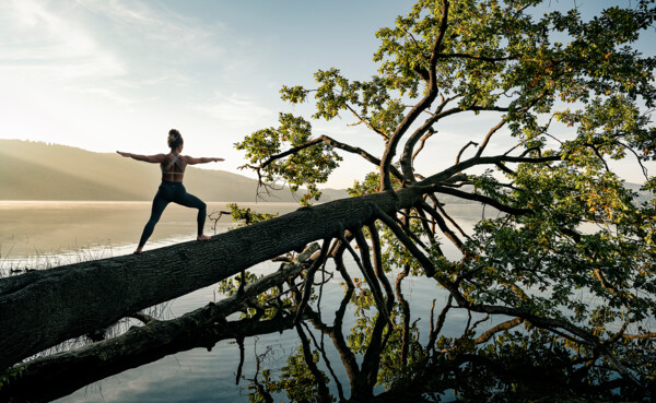 Dame in Kriegerstellung macht Yoga auf einem Baumstamm im Wasser beim Sonnenaufgang bei Maria Laach ©Koblenz-Touristik GmbH, Philip Bruederle