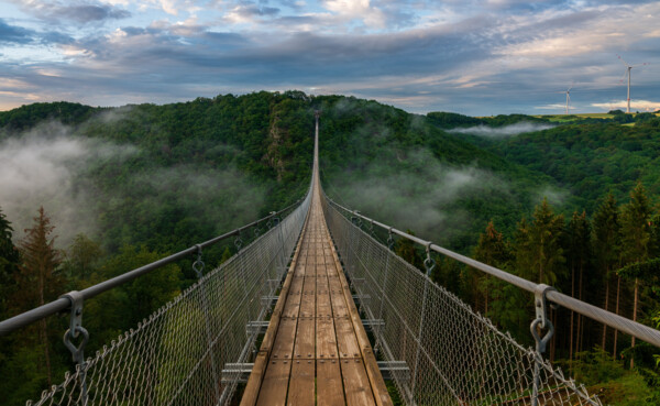 Zicht op een lange hangbrug over een diepe beboste vallei, op de achtergrond bewolkte lucht en mist in de vallei  ©Bernhard - stock.adobe.com