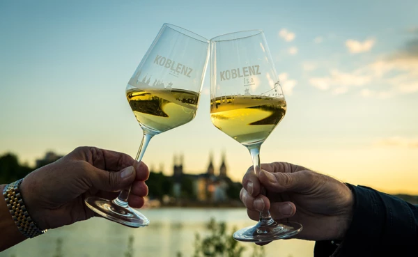 Im Vordergrund werden zwei Weingläser aneinander gehalten, im Hintergrund die Silhouette von Koblenz und die Mosel