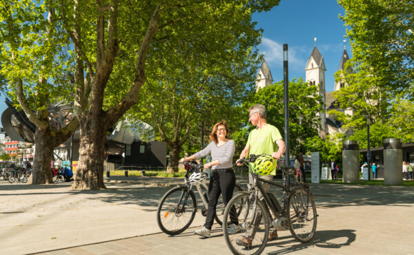 Pärchen mit E-Bikes lächeln. Seilbahn-Talstation und Basilika St. Kastor sind im Hintergrund zu sehen. © Koblenz-Touristik GmbH, Dominik Ketz