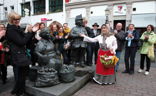 Verkleidete Stadtführerin mit Korb und Gemüse lacht mit einer Führungsgruppe vor Figuren auf dem Münzplatz © Koblenz-Touristik GmbH, Juraschek