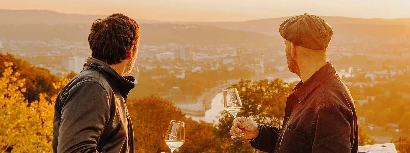 Zwei Männer von der Seite halten Weingläser und blicken auf die Stadt Koblenz beim Sonnenuntergang ©Koblenz-Touristik GmbH, Philip Bruederle