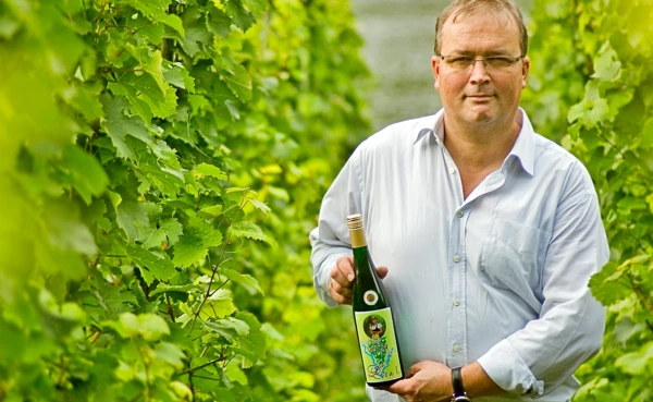 Winzer Rainer Kollman steht mit einer Flasche Wein in seinem Weinberg