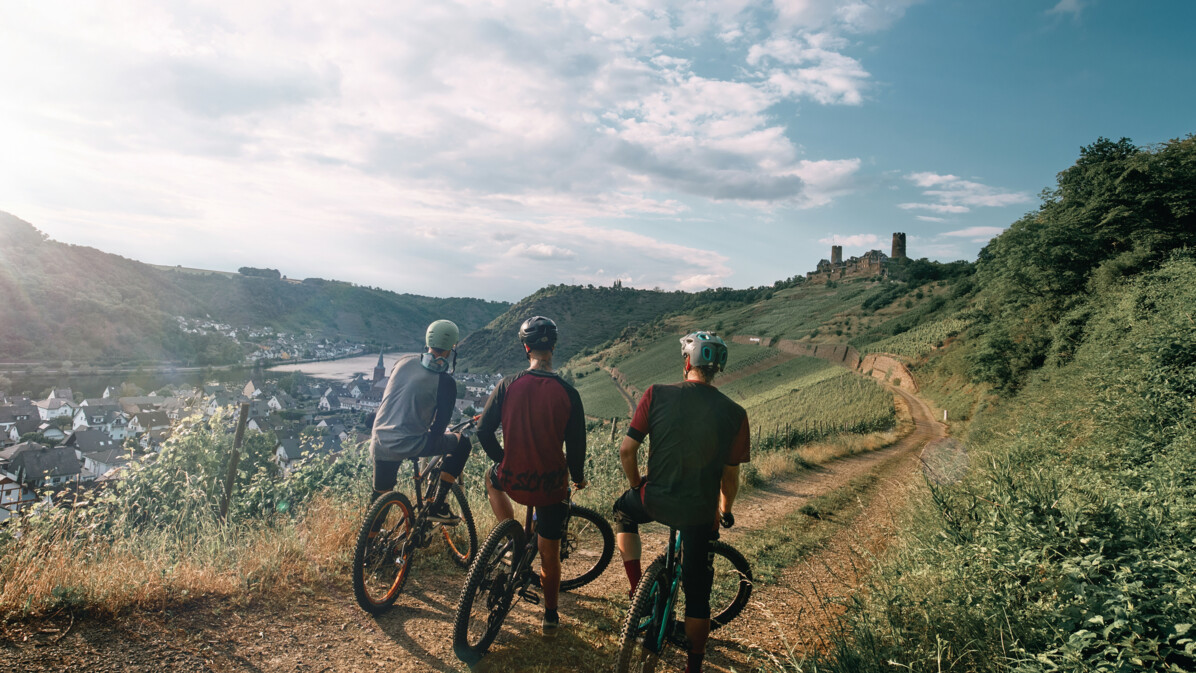 Freundesgruppe auf Mountainbikes auf einem Radweg neben der Mosel mit Burg Thurant im Hintergrund © Philip Bruederle