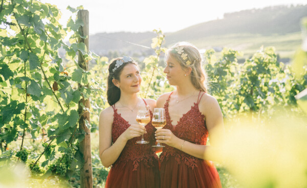 Zwei junge Frauen mit roten Festkleidern, Diadem und Weingläsern, im Hintergrund Weinberge ©Koblenz-Touristik GmbH, Tibor Resch