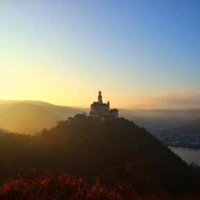 Die Marksburg auf einer Höhe mit Rhein und benebelten Bergen beim Sonnenuntergang ©