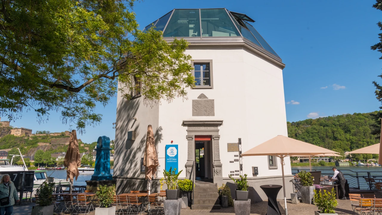 Das Pegelhaus in Koblenz mit Restauranttischen im Vordergrund und dem Rhein und der Festung Ehrenbreitstein im Hintergrund