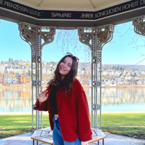 Frau mit rotem Mantel und Sonnenbrille steht im Augusta-Pavillon in den Rheinanlagen, im Hintergrund der Rhein und der Stadtteil Pfaffendorf. ©Kaya Reichert