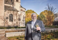 Lächelnder Stadtführer Michael Hörter mit einem Glas Wein in der Hand steht vor dem Basilika St. Kastor im Herbst ©Koblenz-Touristik GmbH, Picture Colada