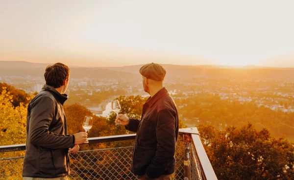 Zwei Männer im Abendlicht mit Weingläsern auf einer Aussichtsplattform mit Blick auf den Zusammenfluss von Rhein und Mosel