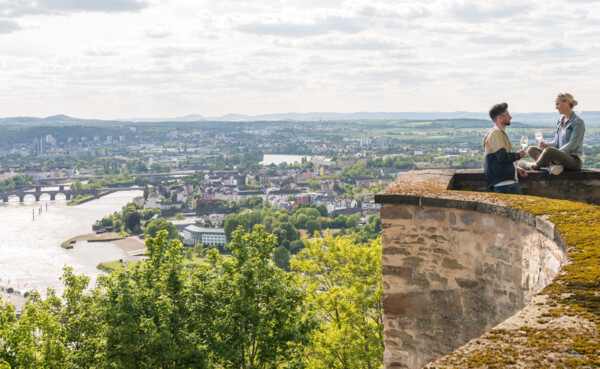 PÃ¤rchen sitzt auf der Mauer der Festung Ehrenbreitstein und hÃ¤lt WeinglÃ¤ser mit Stadt Koblenz im Hintergrund ©Koblenz-Touristik GmbH, Dominik Ketz