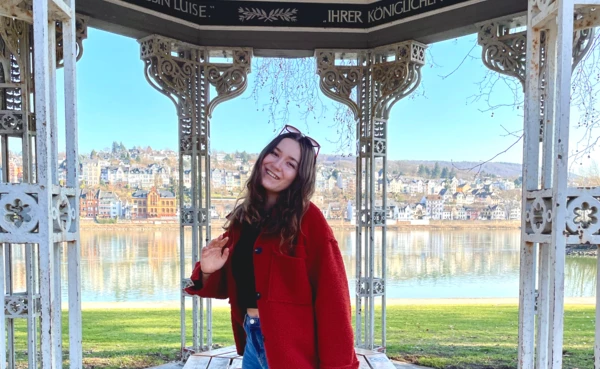 Frau mit rotem Mantel und Sonnenbrille steht im Augusta-Pavillon in den Rheinanlagen, im Hintergrund der Rhein und der Stadtteil Pfaffendorf.