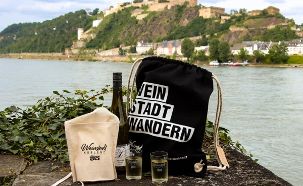 Zwei Mädels sitzen auf der Wiese vor dem preußischen Regierungsgebäude mit einem Glas Wein in der Hand und lachen. Der WeinStadtWander-Beutel steht vor ihnen auf der Wiese. © Koblenz-Touristik, Johannes Bruchhof
