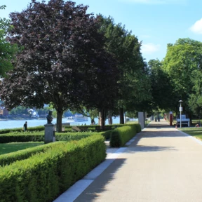Spazier- und Fahrradweg vor dem Weindorf in den Rheinanlagen umgeben von Statuen, Hecken und Bäumen © Koblenz-Touristik GmbH