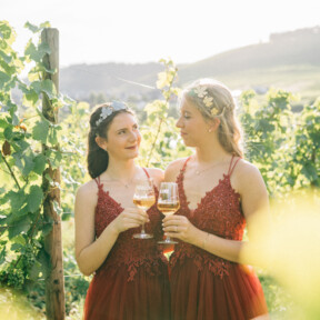 Zwei junge Frauen mit roten Festkleidern, Diadem und Weingläsern, im Hintergrund Weinberge ©Koblenz-Touristik GmbH, Tibor Resch