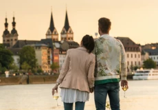 Pärchen mit Wein in der Hand lehnen an einander beim Zuschauen der Stadtsilhouette beim Sonnenuntergang.
