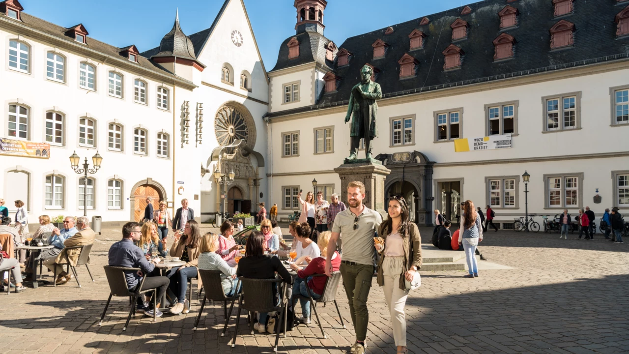 Pärchen von Vorne geht und Menschengruppen sitzen an Cafétischen auf dem Jesuitenplatz in der Koblenzer Altstadt
