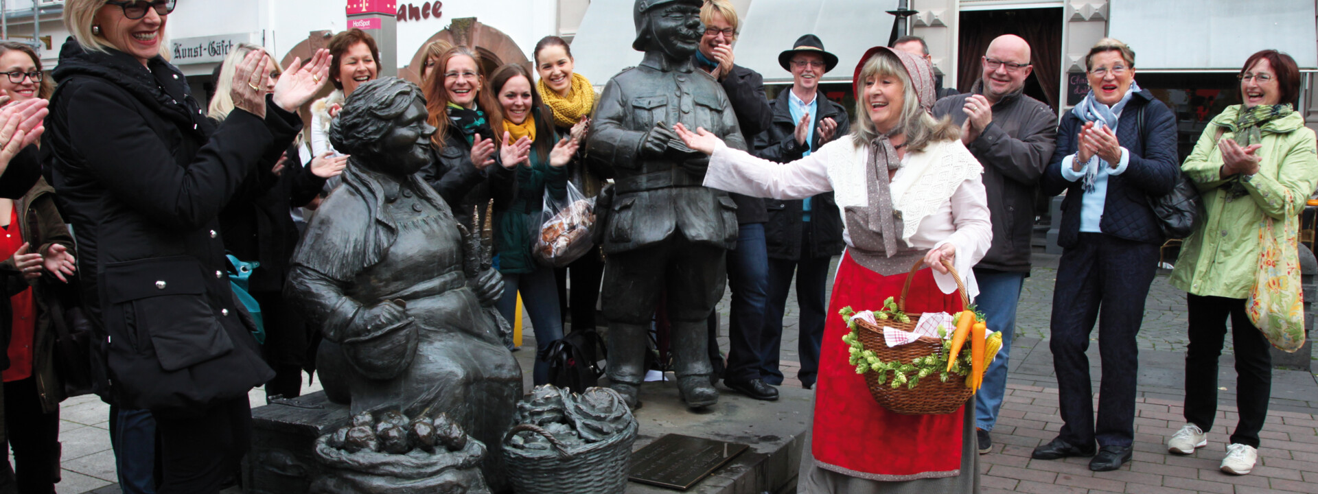 Verkleidete Stadtführerin mit Korb und Gemüse lacht mit einer Führungsgruppe vor Figuren auf dem Münzplatz ©Koblenz-Touristik GmbH, Juraschek