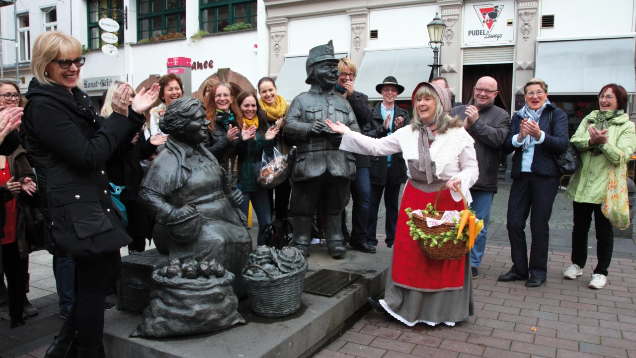 Verkleidete Stadtführerin mit Korb und Gemüse lacht mit einer Führungsgruppe vor Figuren auf dem Münzplatz