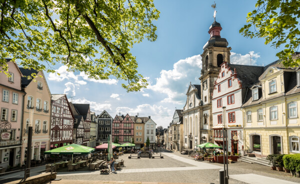 Hachenburg marketplace with tables and umbrellas in front of restaurants, surrounded by colorful half-timbered houses and a church, in the foreground branches, in the background blue sky ©Tourist-Info Hachenburger Westerwald, Dominik Ketz