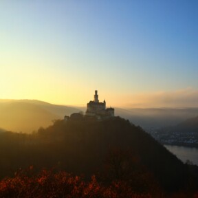 Die Marksburg auf einer Höhe mit Rhein und benebelten Bergen beim Sonnenuntergang © 
