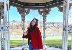 Frau mit rotem Mantel und Sonnenbrille steht im Augusta-Pavillon in den Rheinanlagen, im Hintergrund der Rhein und der Stadtteil Pfaffendorf. ©Kaya Reichert